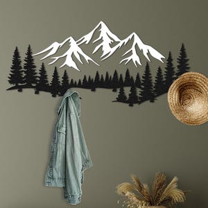 Puede incluir: Una pieza de arte mural de metal negro con montañas y bosque está montada en una pared verde. Una chaqueta vaquera cuelga debajo. Un sombrero tejido y un mueble de madera con decoración completan la escena.