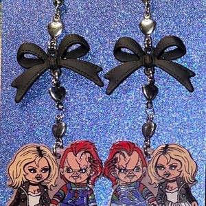 May include: A pair of dangle earrings featuring black bows, silver chains with heart charms, and a charm of a couple from the horror movie Child's Play. The couple is made up of Chucky and Tiffany.
