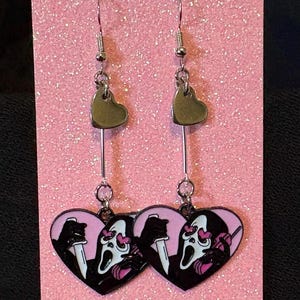May include: A pair of silver drop earrings. Each earring has a small silver heart at the top, linked to a heart-shaped charm with a black and pink design of a ghost face holding a knife. The earrings are displayed on a pink glittery surface.