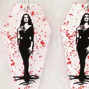May include: A pair of coffin-shaped earrings. Each earring displays a black silhouette of a woman in a long dress, with red splatter on a white background. The earrings are suspended from silver hoops.