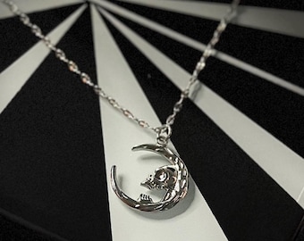 May include: Silver necklace with a crescent moon pendant, featuring a skull design. The chain is delicate. The background has a black and white geometric pattern.
