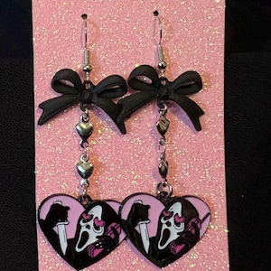 May include: Dangle earrings with heart-shaped pendants featuring a Scream mask design. The earrings have black bows, silver-toned chains with heart charms, and are displayed on a pink glittery surface. A spooky accessory.