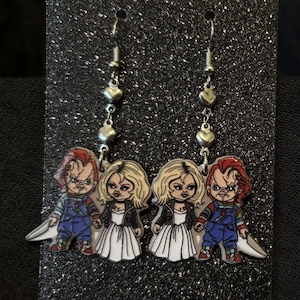 May include: A pair of dangle earrings featuring Chucky and Tiffany Valentine characters. The earrings have silver-toned hooks and heart-shaped accents. The Chucky figures wear blue overalls, while the Tiffany figures wear white dresses and black jackets.