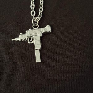 May include: Silver-toned metal pendant necklace featuring a detailed miniature Uzi submachine gun charm. The gun charm is suspended from a linked chain. The background is a dark, solid color.