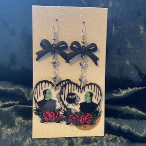 May include: Dangle earrings with black bows, silver heart charms, and heart-shaped pendants. The pendants feature Frankenstein's monster and bride, red roses, and a black and white striped background.