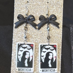 May include: Dangle earrings with black bows, silver chains, and rectangular pendants. The pendants show a Morticia Addams portrait with bats and the name "MORTICIA". The earrings are displayed on a glittery gold surface, ideal for fans of the Addams Family.