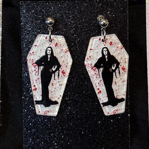 May include: A pair of coffin-shaped earrings. Each earring features a black silhouette of a woman in a long dress, set against a white background with red splatter. They are attached to silver-coloured ball studs.