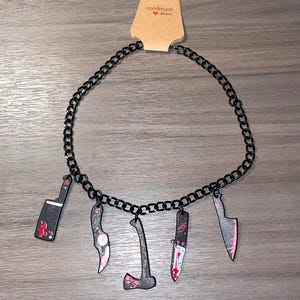 May include: A black chain necklace with five charms: a butcher knife, a cleaver, an axe, and two knives. The charms are painted with red and black to look like blood splatters. The tag on the necklace reads "Handmade with love."