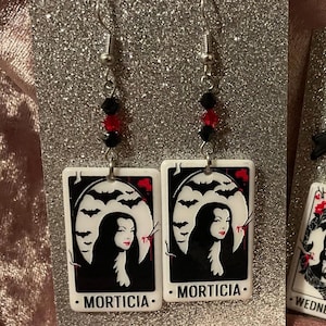 May include: Dangle earrings with silver hooks, featuring Morticia Addams from The Addams Family. Each earring has a rectangular charm with a black and white illustration of Morticia, with a bat silhouette and the word "MORTICIA". Red and black beads.