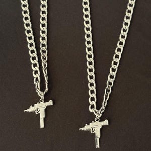 May include: Two silver-colored necklaces with a gun pendant. The chains are thick and linked. The pendants are detailed replicas of a gun. The background is a solid, dark brown color.