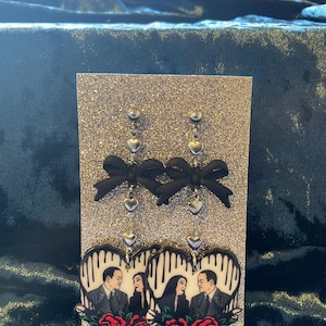May include: A pair of dangle earrings featuring a black and white image of a couple in heart-shaped frames. The earrings have a silver chain with black bows and silver heart charms.
