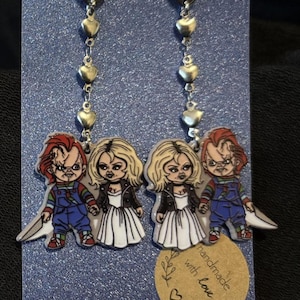 May include: Dangle earrings featuring Chucky and Tiffany from a horror film. The earrings have silver heart accents and are set against a blue glitter background. A small tag reads "handmade with love."