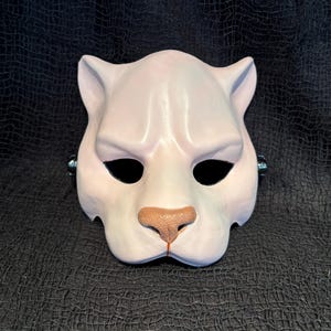 以下が含まれることがあります： 黒い目の穴と茶色の鼻が付いた白い猫のマスク。マスクは滑らかでマットな仕上がりで、顔を覆うように設計されています。マスクには小さな耳があり、耐久性のある素材で作られている可能性があります。マスクはコスチュームパーティーや演劇に適しています。