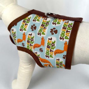 May include: A dog harness with a light blue background featuring orange squirrels, paw prints, and the words "Squirrel Patrol" in white and green. The harness has brown trim and a silver metal ring.