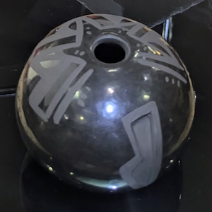 May include: A spherical, black ceramic pot with a shiny surface. It features etched geometric designs in grey. The pot has a central hole and a curved, rectangular handle-like feature. The pot is likely hand-crafted.