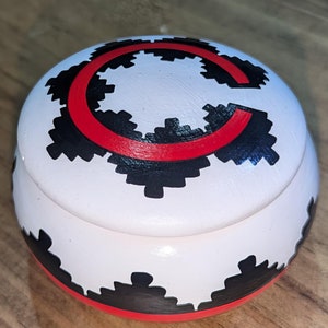 May include: A round, white wooden box with a lid. The box is decorated with a black and red design, including a large red "C" and black geometric shapes. The box sits on a wooden surface.