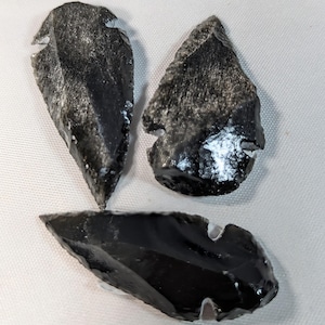 May include: Three black obsidian arrowheads with a sharp point and a chipped edge. The arrowheads are on a white surface.