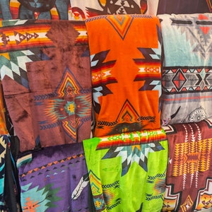 Puede incluir: Una exhibici&oacute;n de mantas plegadas con dise&ntilde;os de inspiraci&oacute;n suroeste. Las mantas presentan dise&ntilde;os geom&eacute;tricos en varios colores, incluyendo naranja, marr&oacute;n, morado y verde. Algunas mantas tienen motivos tribales y dise&ntilde;os de plumas. Las mantas est&aacute;n hechas de un material suave y afelpado.