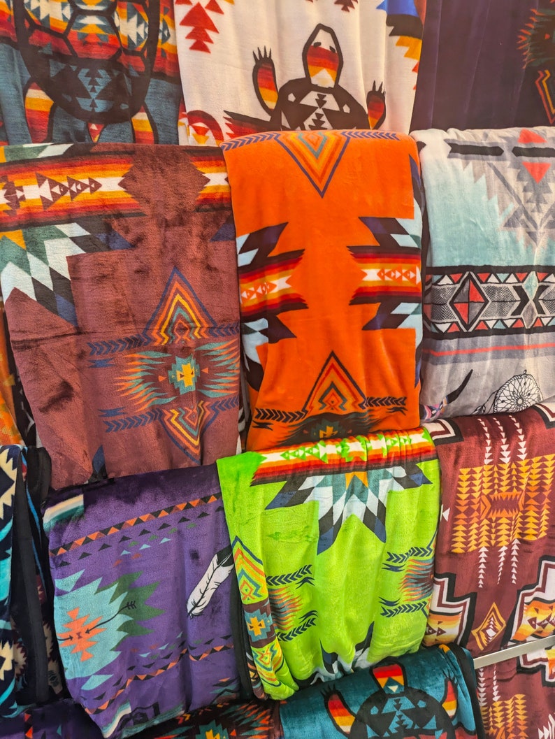 Puede incluir: Una exhibici&oacute;n de mantas plegadas con dise&ntilde;os de inspiraci&oacute;n suroeste. Las mantas presentan dise&ntilde;os geom&eacute;tricos en varios colores, incluyendo naranja, marr&oacute;n, morado y verde. Algunas mantas tienen motivos tribales y dise&ntilde;os de plumas. Las mantas est&aacute;n hechas de un material suave y afelpado.
