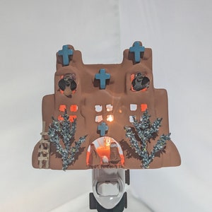 Puede incluir: Una luz de noche de cerámica con forma de iglesia de adobe marrón con cruces azules y campanas. La luz de noche tiene un brillo cálido que proviene de la ventana.