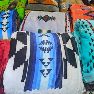 Puede incluir: Una colecci&oacute;n de mantas plegadas y enrolladas con varios patrones geom&eacute;tricos. Las mantas presentan una gama de colores, incluyendo azul, negro, blanco, naranja y verde. Los dise&ntilde;os incluyen motivos tribales e inspirados en el suroeste.