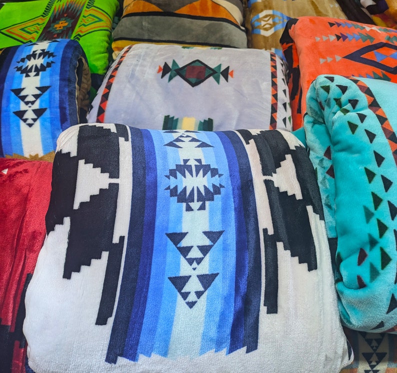 Puede incluir: Una colecci&oacute;n de mantas plegadas y enrolladas con varios patrones geom&eacute;tricos. Las mantas presentan una gama de colores, incluyendo azul, negro, blanco, naranja y verde. Los dise&ntilde;os incluyen motivos tribales e inspirados en el suroeste.
