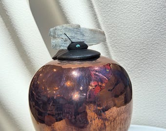 May include: A copper-colored, round vase with a textured surface. It is topped with a stack of black stones, a small turquoise accent, and a light blue stone. The vase reflects light, creating a warm, inviting aesthetic.