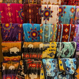 Puede incluir: Una exhibici&oacute;n de mantas plegadas con varios dise&ntilde;os de inspiraci&oacute;n nativa americana. Las mantas presentan patrones geom&eacute;tricos, explosiones de estrellas e im&aacute;genes de bisontes en tonos rojos, azules, morados y amarillos.