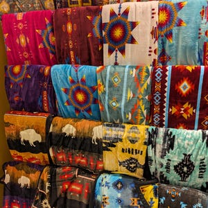 Puede incluir: Una exhibici&oacute;n de mantas plegadas con varios dise&ntilde;os de inspiraci&oacute;n nativa americana. Las mantas presentan patrones geom&eacute;tricos, dise&ntilde;os de estrellas y siluetas de b&uacute;falos en colores como rojo, azul, amarillo y marr&oacute;n. Las mantas est&aacute;n apiladas en estantes.