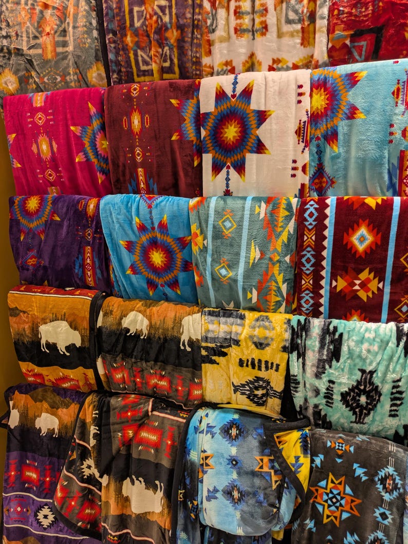 Puede incluir: Una exhibici&oacute;n de mantas plegadas con varios dise&ntilde;os de inspiraci&oacute;n nativa americana. Las mantas presentan patrones geom&eacute;tricos, dise&ntilde;os de estrellas y siluetas de b&uacute;falos en colores como rojo, azul, amarillo y marr&oacute;n. Las mantas est&aacute;n apiladas en estantes.