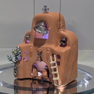 May include: A small, terracotta-colored model of a church with a cross on top. The church has a bell tower with a bell on each side, and a ladder on the side. There is a small tree with white flowers in front of the church.