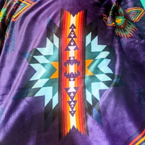 Puede incluir: Primer plano de un textil morado con un patr&oacute;n geom&eacute;trico. El dise&ntilde;o presenta un motivo central de estrella con tri&aacute;ngulos y rayas en tonos de azul, naranja, rojo y blanco. La tela tiene una textura suave y aterciopelada.