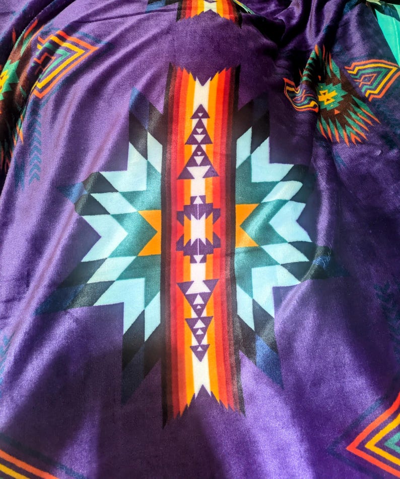 Puede incluir: Primer plano de un textil morado con un patr&oacute;n geom&eacute;trico. El dise&ntilde;o presenta un motivo central de estrella con tri&aacute;ngulos y rayas en tonos de azul, naranja, rojo y blanco. La tela tiene una textura suave y aterciopelada.