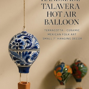 Puede incluir: Globo aerostático Talavera hecho a mano, una decoración colgante de 18 cm. El globo de cerámica presenta un diseño floral azul y blanco. El globo está suspendido por una cuerda y tiene una cesta de terracota. El texto en la imagen dice "ALL TRIBES TREASURES".