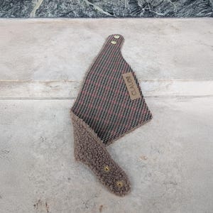 Könnte beinhalten: Ein Hunde-Bandana mit einem rot-schwarz-weiß karierten Muster und einem weichen, braunen, flauschigen Futter. Das Bandana hat ein kleines rechteckiges Etikett mit der Aufschrift "CLAUDE". Es wird mit goldfarbenen Druckknöpfen befestigt.