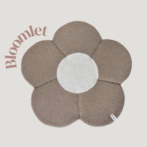 Può includere: Tappeto a forma di fiore con petali marroni e centro bianco. Il tappeto ha un aspetto morbido e strutturato. La parola "bloomlet" è stampata sul tappeto. Una piccola etichetta è attaccata al tappeto.