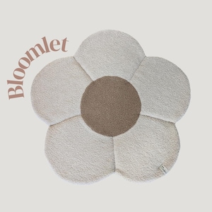 Può includere: Un cuscino da pavimento a forma di fiore con un design a petali bianco sporco e strutturato e un centro marrone. La parola "bloomlet" è stampata sul lato superiore sinistro del cuscino. Il cuscino è progettato per il comfort e la decorazione.