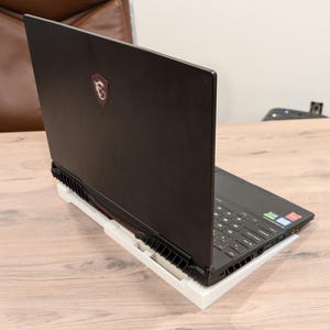 May include: A black laptop computer with a red logo on the back is sitting on a white stand on a wooden table.