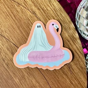 May include: A sticker featuring a ghost and a pink flamingo pool float. The ghost is draped over the flamingo, floating on a blue and orange water background. The sticker is on a wooden surface, with disco balls and a glittery bone in the background.