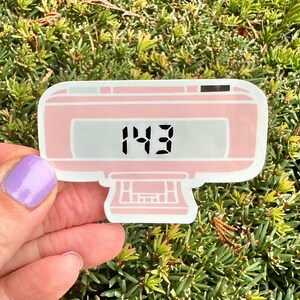 May include: Pink digital alarm clock sticker with a white display showing the number 143. The sticker is on a green background.
