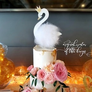 Peut inclure: Un gâteau blanc à deux niveaux décoré de roses roses, de verdure et d'accents dorés. Un cake topper cygne blanc avec une couronne dorée et des plumes duveteuses est posé sur le gâteau. Le texte "original creator of this design" est visible.