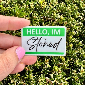 May include: A white sticker with green trim and the text "HELLO, IM STONED" in black cursive writing.