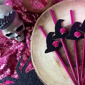 Puede incluir: Conjunto de seis pajitas de bruja de color rosa y negro con una rosa rosa en cada sombrero. Las pajitas están sobre una rodaja de madera.