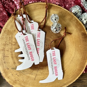 May include: A set of five white boot-shaped tags with brown string. Each tag has the text "The Last Rodeo" and "Lauren's Bachelorette" printed on it. The tags are on a wooden surface with silver disco balls.