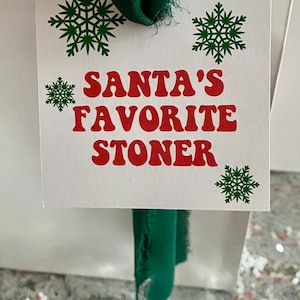 May include: White card with green snowflakes and red text that reads "Santa's Favorite Stoner". A green fabric ribbon is attached to the top of the card.