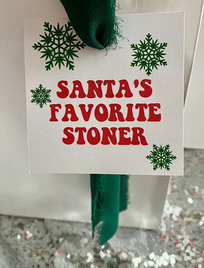 May include: White card with green snowflakes and red text that reads "Santa's Favorite Stoner". A green fabric ribbon is attached to the top of the card.