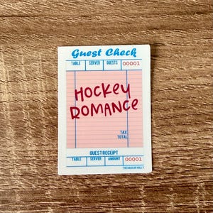 May include: A sticker resembling a guest check with the words "Guest Check" in blue and "Hockey Romance" in red. The sticker has a pink background and the text "The Haus of Holly" at the bottom.