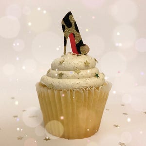 May include: A cupcake with white frosting, decorated with gold star sprinkles and a high-heeled shoe topper. The shoe is black with gold glitter accents and a red sole. The cupcake sits in a light brown paper liner.