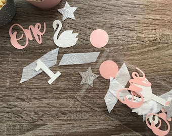 May include: Pink and white confetti with a silver glitter star, a white swan, and the word "one" in pink. The confetti is in a clear plastic bag.