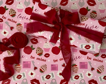 May include: A rectangular gift box wrapped in light pink paper featuring a repeating pattern of red roses, chocolate-covered strawberries, pink envelopes, keys, and tickets with lips. A dark red frayed ribbon is tied in a bow on top.